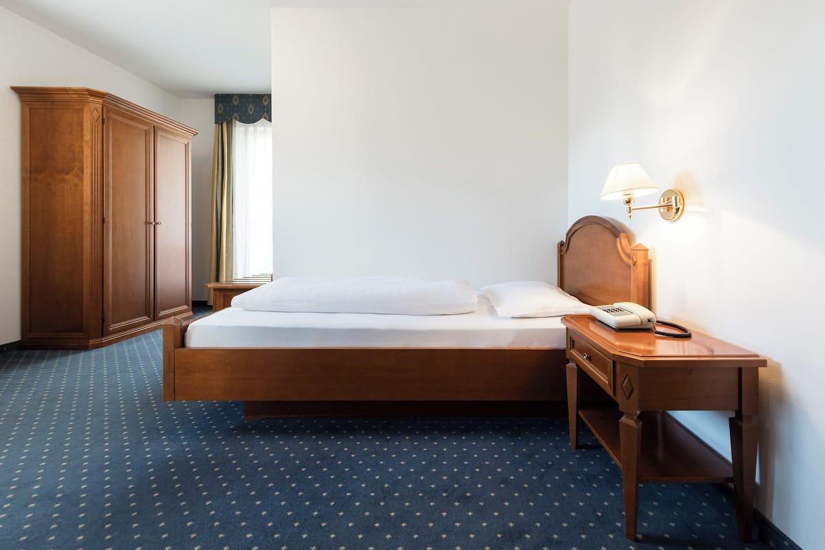 Una habitación de hotel con cama, un armario de madera, una mesita de noche con lámpara y un teléfono.