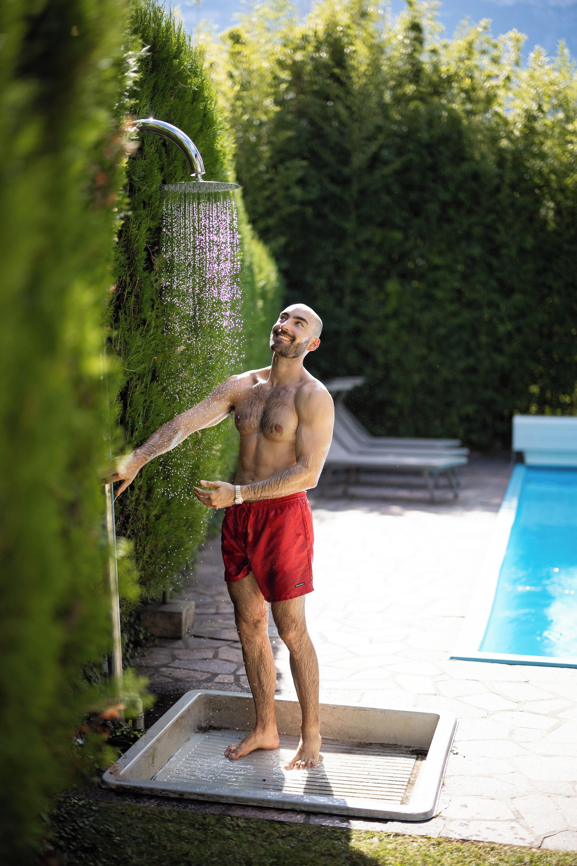 Un hombre de pie sobre una plataforma metálica junto a una piscina, con un jardín y un seto al fondo.
