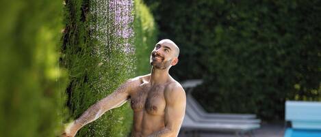 Un hombre de pie sobre una plataforma metálica junto a una piscina, con un jardín y un seto al fondo.
