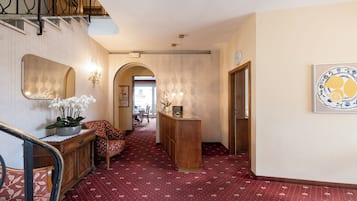 A hotel lobby with a red patterned carpet, a reception desk, a wooden chair, a large mirror, and a wall-mounted clock.