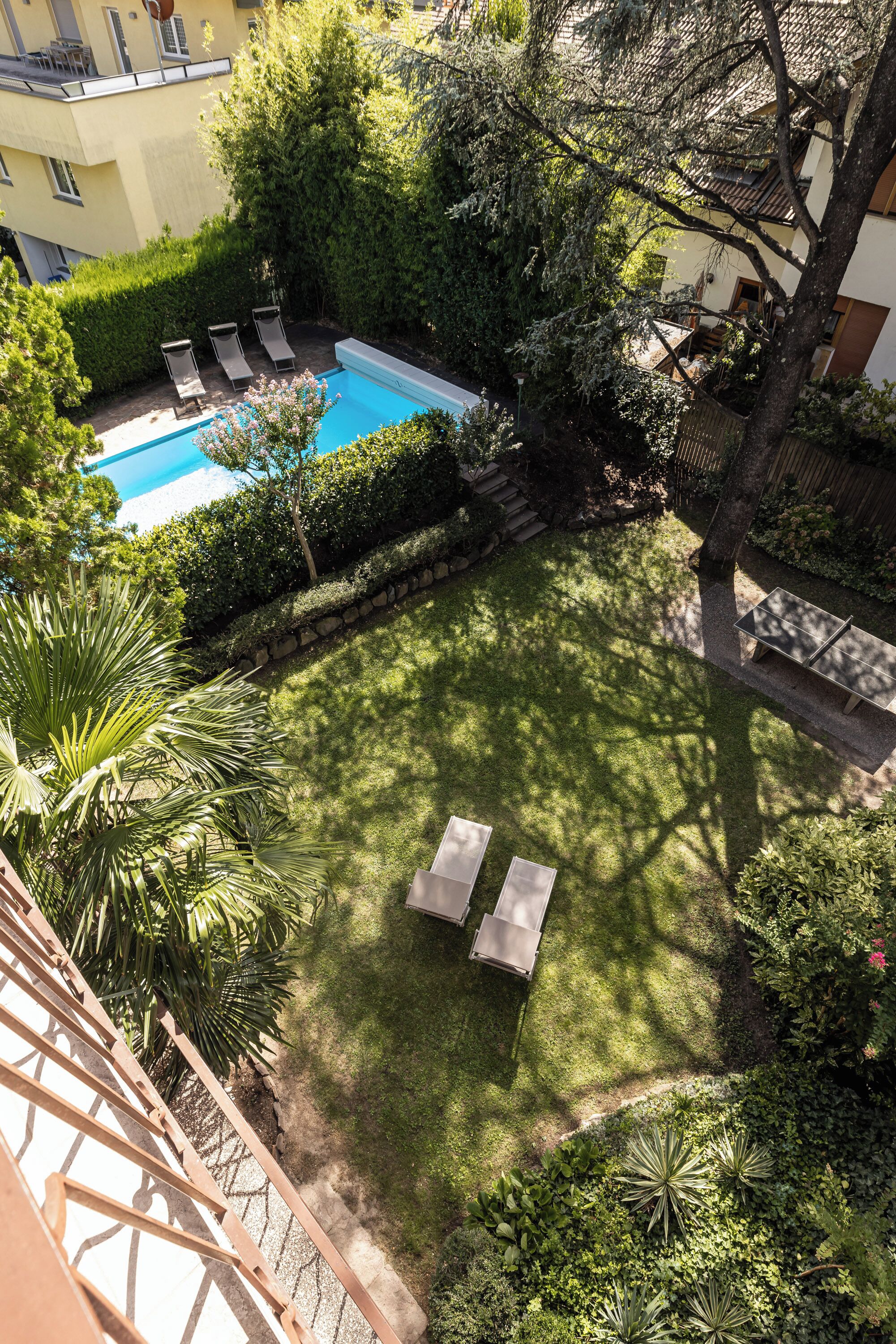 A rooftop pool area with lounge chairs and a small table, surrounded by lush greenery and a clear blue pool.