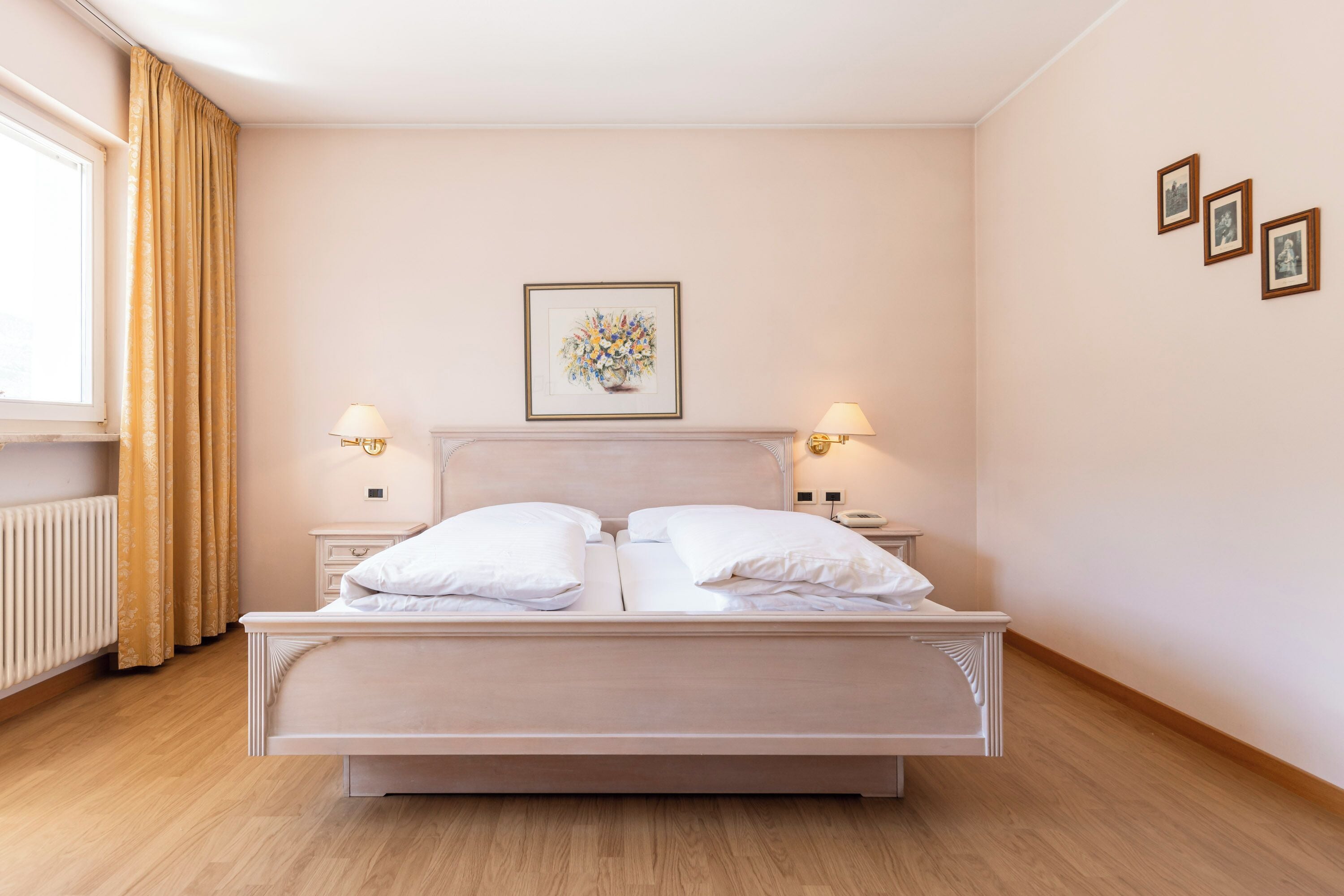 Una cama bien hecha con sábanas blancas en una habitación con suelo de madera clara, un radiador y una ventana con cortinas.