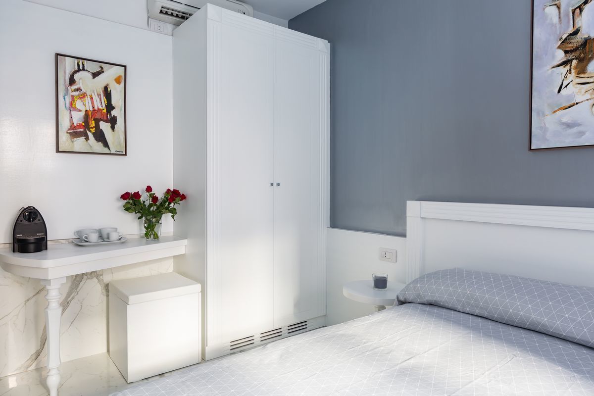 A bedroom with a bed, a white wardrobe, a bedside table with a cup and flowers, and a painting on the wall.