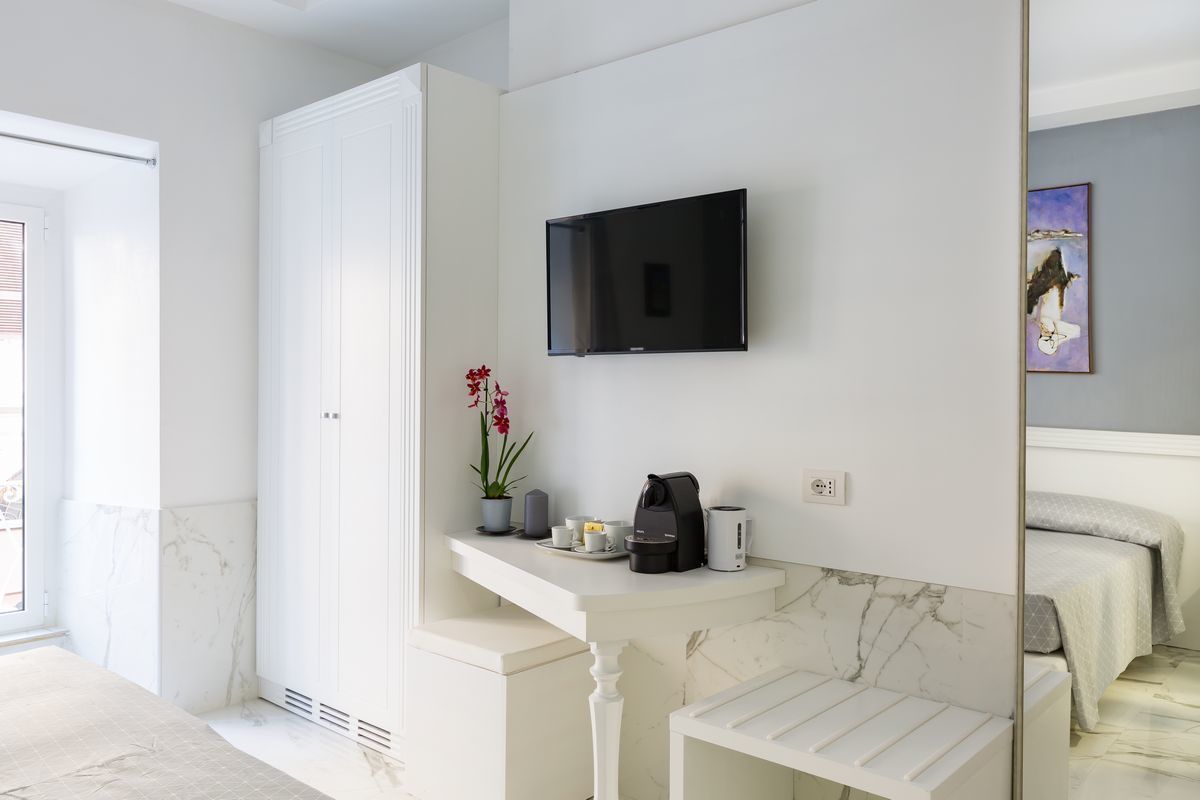 Una habitació d'hotel moderna amb un sòl de marbre, un escriptori blanc amb una cafetera, un televisor de pantalla plana i un llit amb una colcha gris.