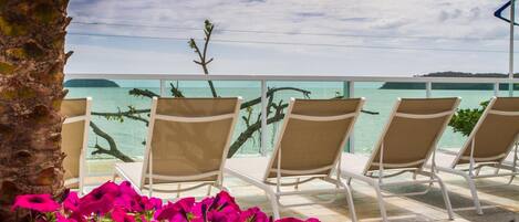 A terrace with lounge chairs and vibrant pink flowers, overlooking a calm sea and distant land.