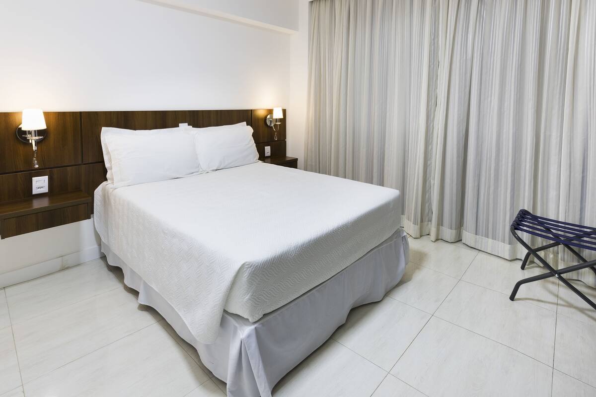 Una cama bien hecha con cubrecama y almohadas blancas, cabecera de madera, dos lámparas de noche y una silla a la derecha.