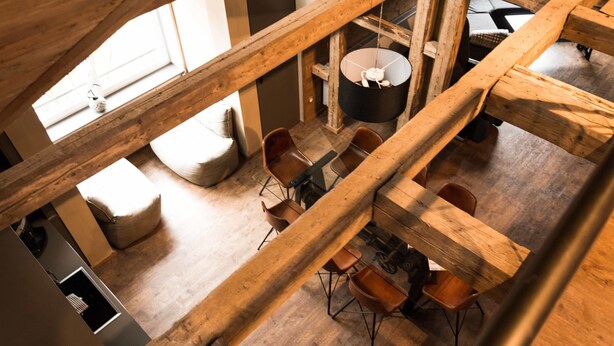 A high-ceilinged wooden dining area with a central hanging light fixture, multiple wooden beams, and a view of the kitchen through a window. - Die Hütten 550 üNN (Braunlage)