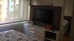 Une chambre à coucher avec un lit, un téléviseur posé sur un meuble, une commode et une fenêtre avec des stores.