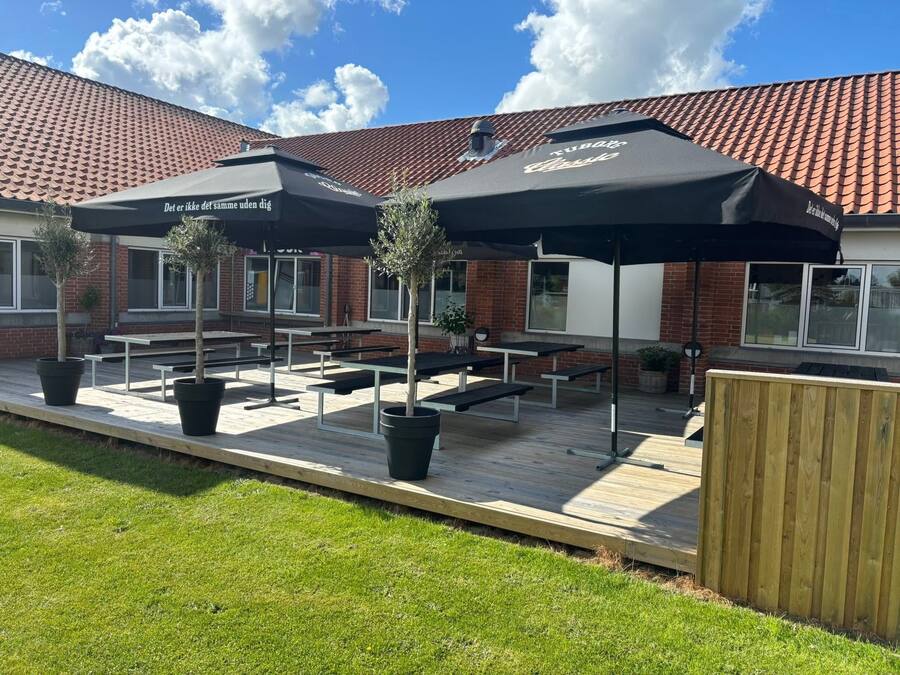 An outdoor seating area with tables and umbrellas, a brick building in the background, and a wooden fence.