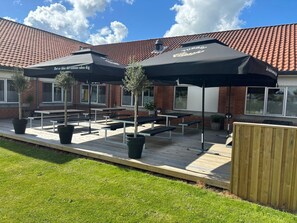An outdoor seating area with tables and umbrellas, a brick building in the background, and a wooden fence.