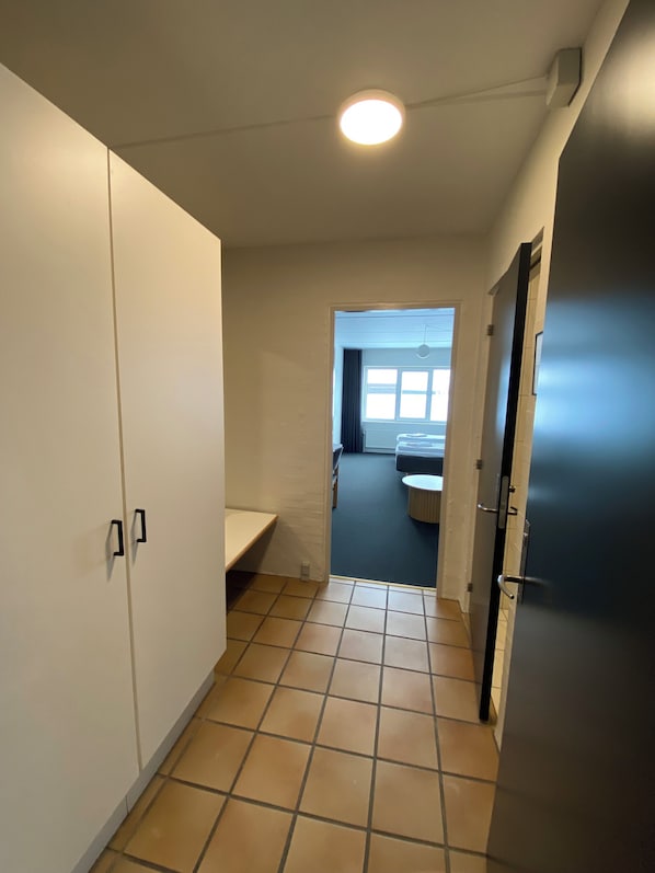 Un pasillo estrecho con suelo de baldosas, un armario blanco, un pequeño banco y una puerta que da a una habitación con ventana y mesa redonda.