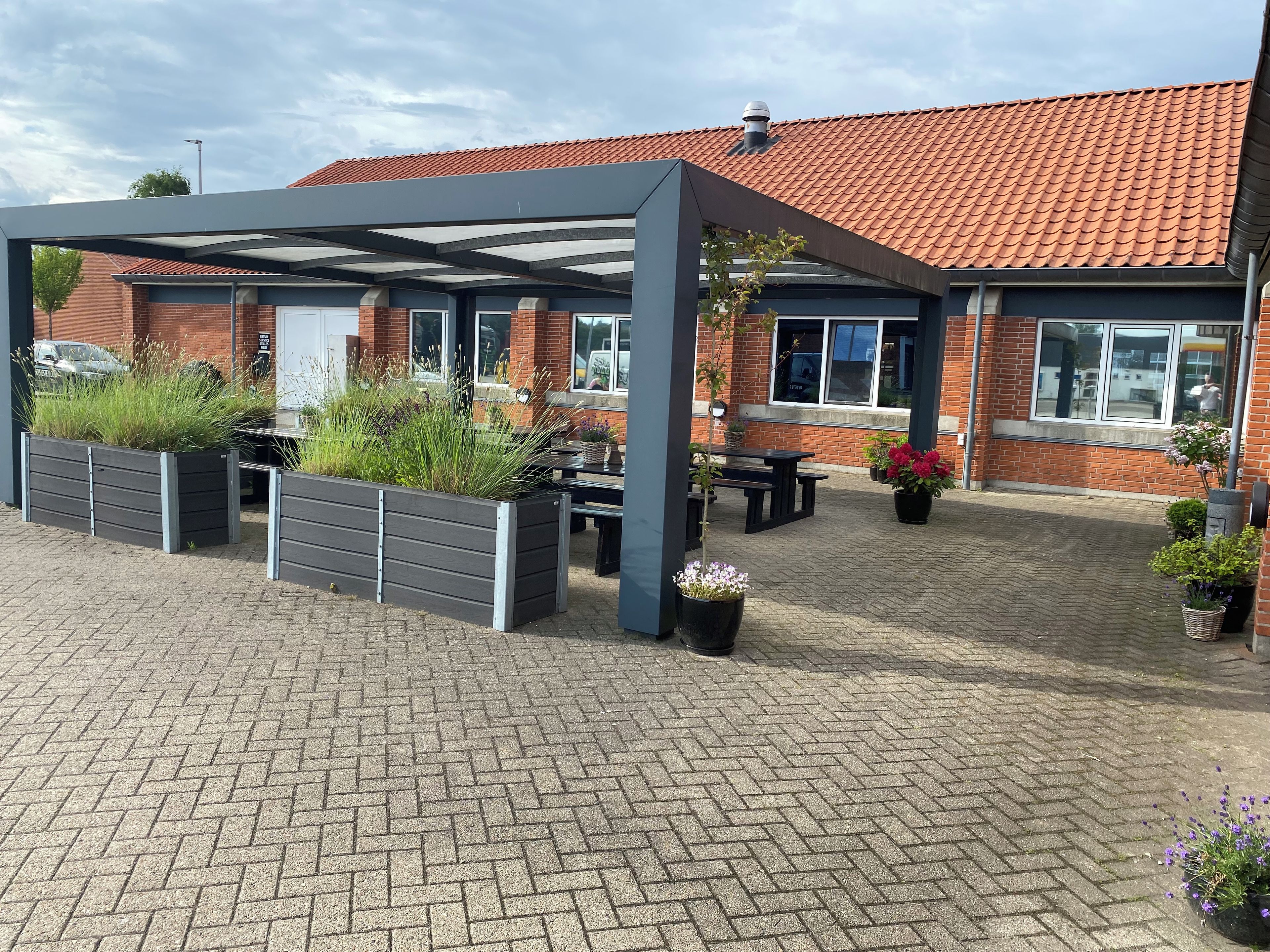 A covered outdoor seating area with planters and a brick building in the background.