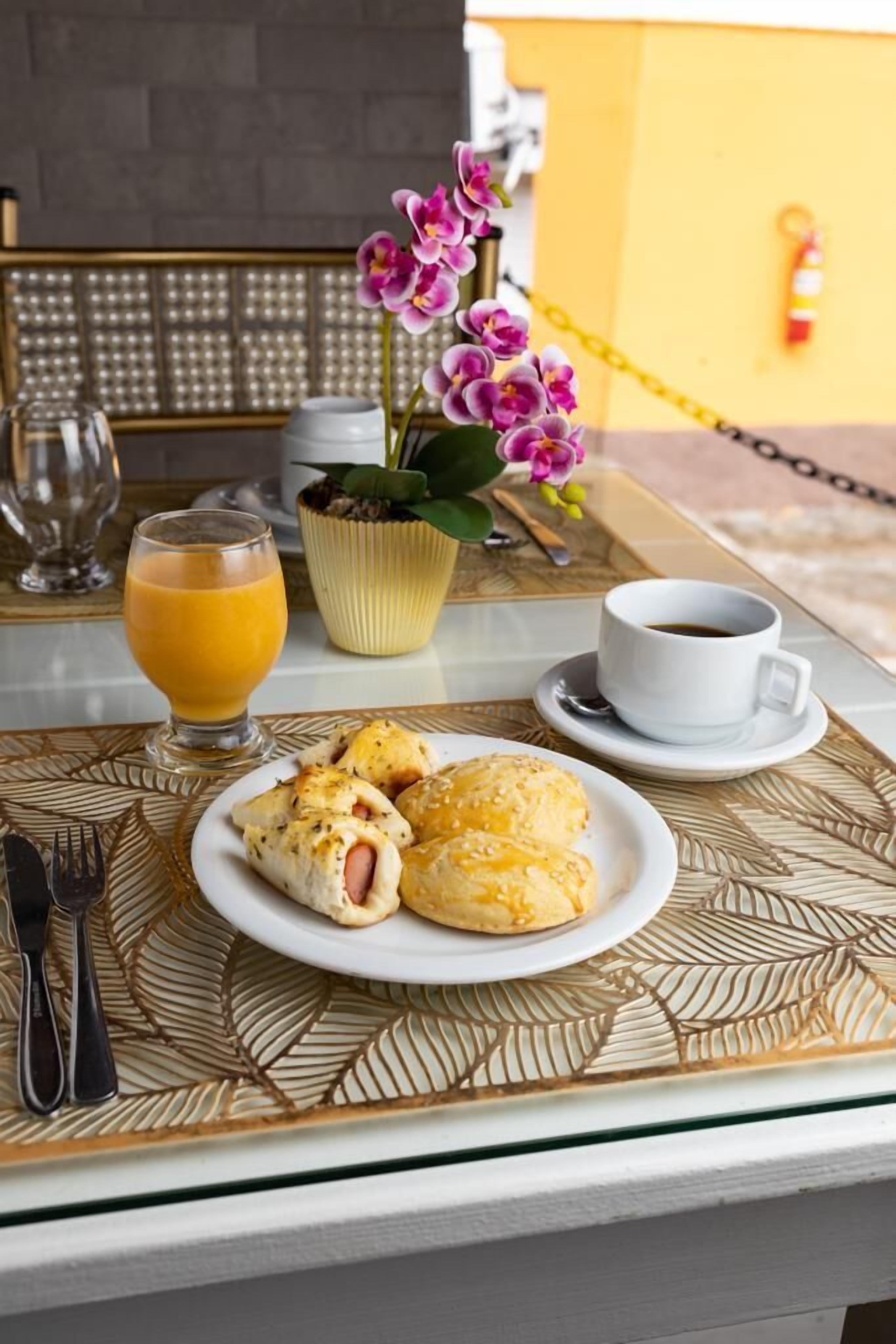 Une table mise pour le petit-déjeuner, avec une assiette de viennoiseries, un verre de jus d’orange et une tasse de café, le tout posé sur une nappe décorative.