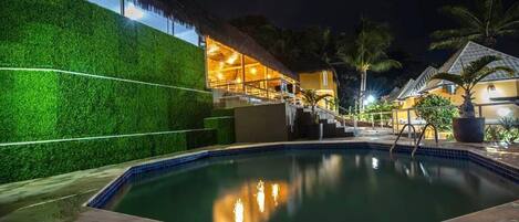 A swimming pool with a thatched roof structure, surrounded by a lush green hedge and palm trees.