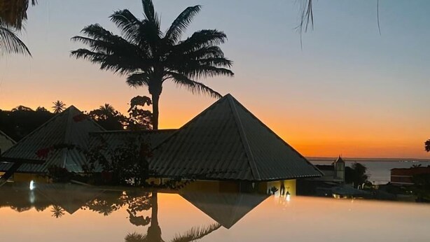 Una noche tranquila en un resort, con un cielo despejado, una piscina serena que refleja palmeras y una estructura con techo de paja.