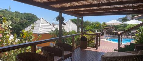 A wooden deck with wicker furniture overlooking a swimming pool and a view of buildings with tiled roofs.