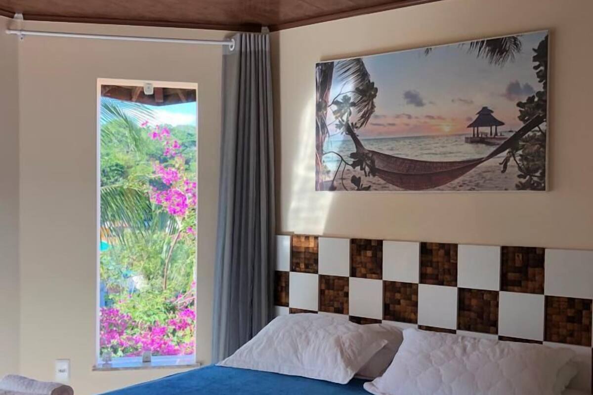 A bedroom with a bed, a window with a view of greenery, and a painting of a beach scene.