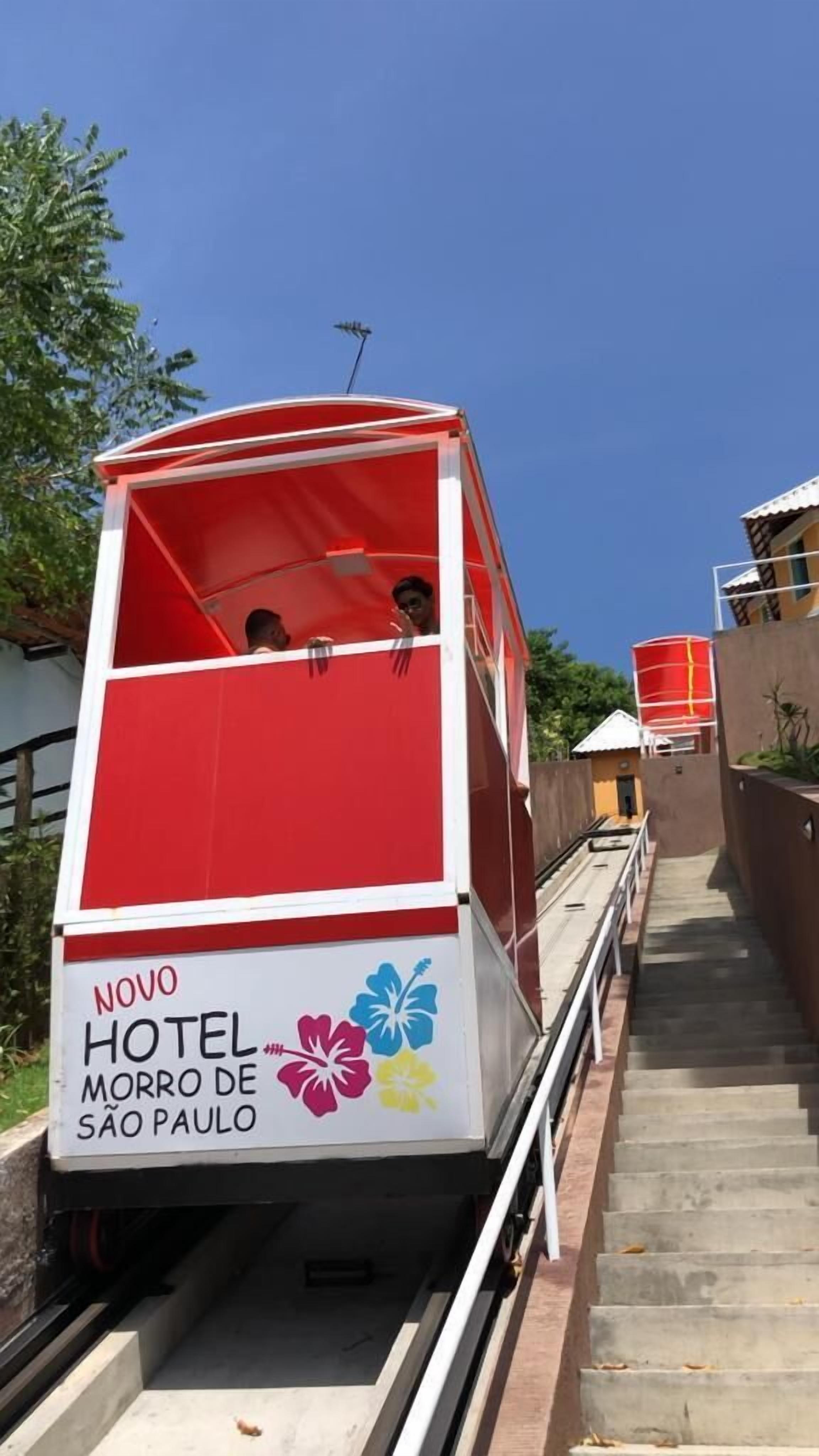 A red and white cable car with "NOVO HOTEL MORRO DE SÃO PAULO" written on the side, moving along a track.
