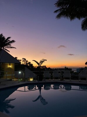 Une vue sur la piscine au crépuscule, avec des chaises longues et un bâtiment en arrière-plan.
