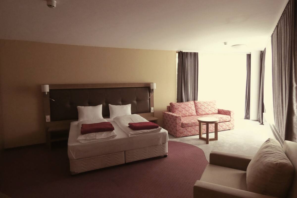 Une chambre d’hôtel avec un lit, un canapé, une petite table et une fenêtre avec des rideaux.