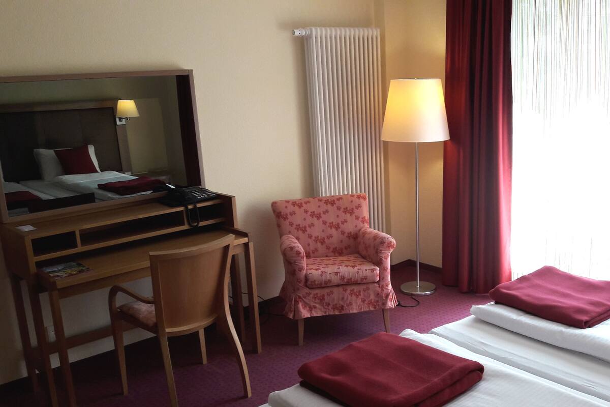 Une chambre d’hôtel avec un lit, un bureau, une chaise, une lampe et une fenêtre avec des rideaux.
