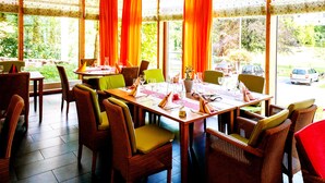 Un coin repas avec des chaises en osier et une table dressée pour le repas, de grandes fenêtres avec des rideaux et une vue sur l’extérieur.