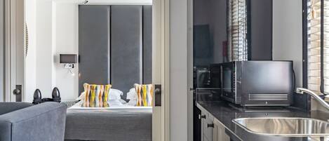 A modern hotel room with a bed, a sofa, a kitchen area with a microwave, and a sink.