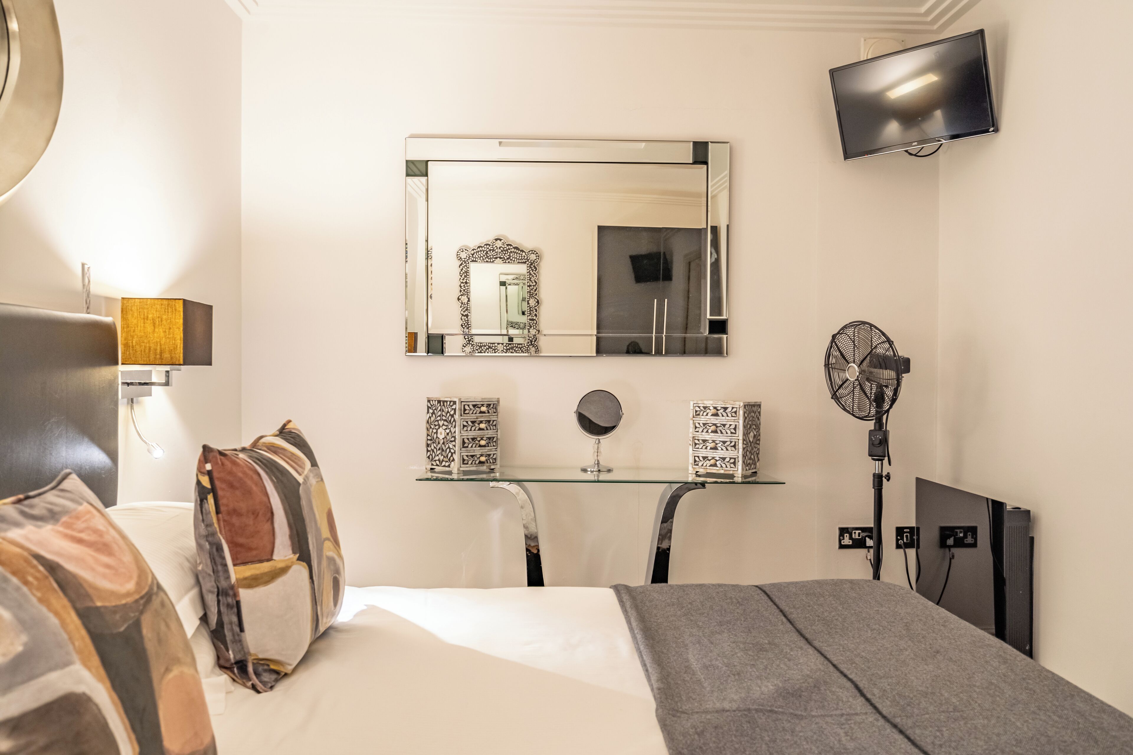 A hotel room with a bed, a desk with a mirror, a television, and a fan.