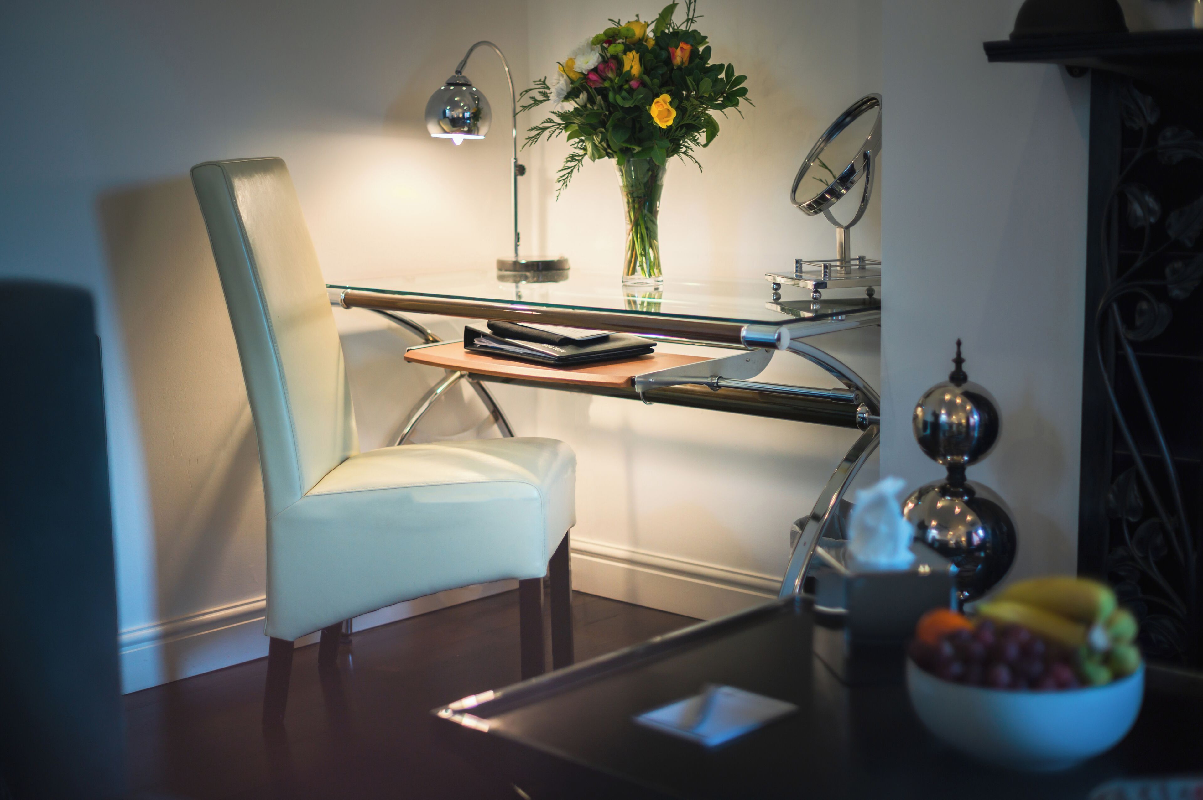 A modern office chair, a desk with a lamp, a vase with flowers, and a bowl of fruit.