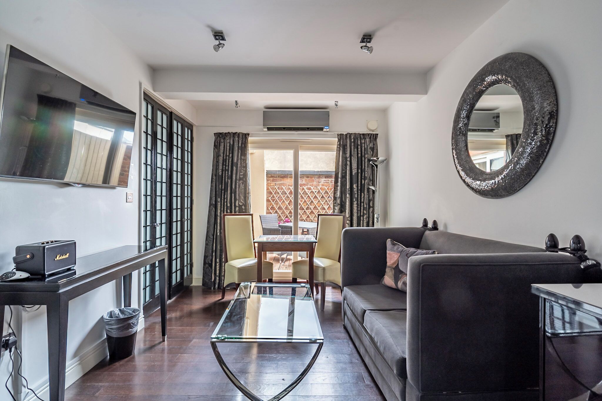 A modern living room with a glass coffee table, a sofa, a TV mounted on the wall, and a large mirror.