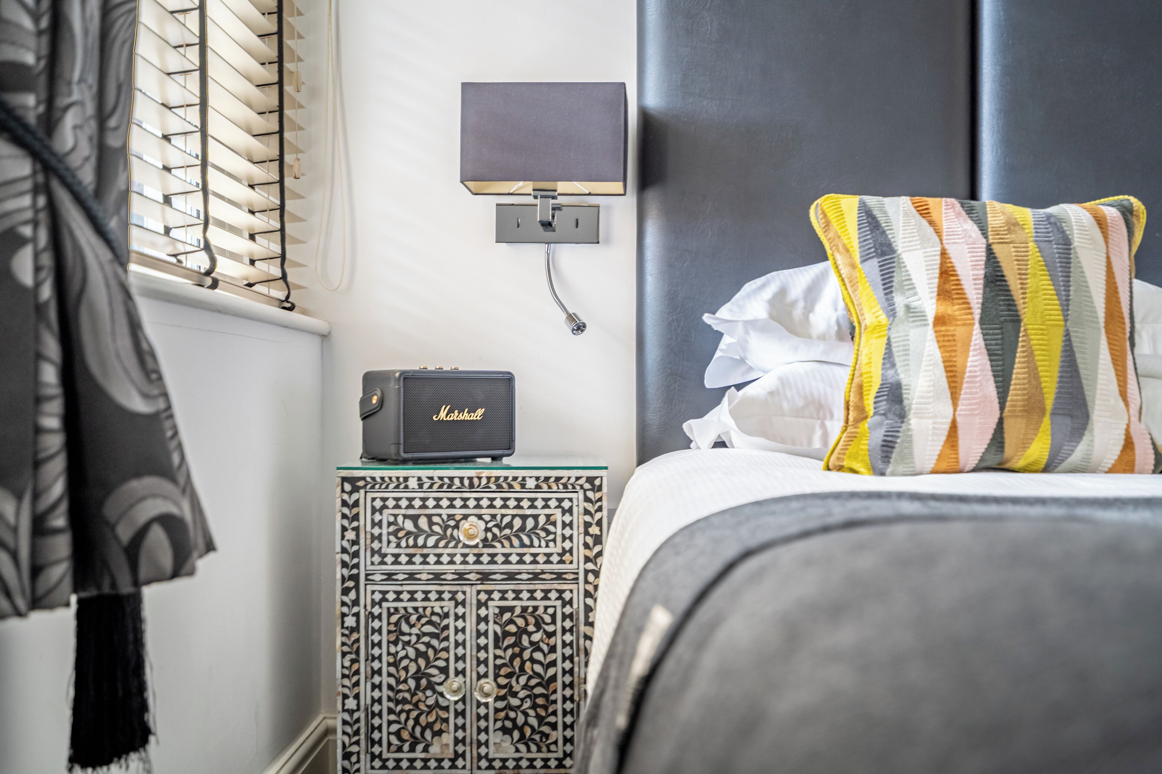 A neatly made bed with a striped pillow, a bedside table with a radio, and a wall-mounted lamp.