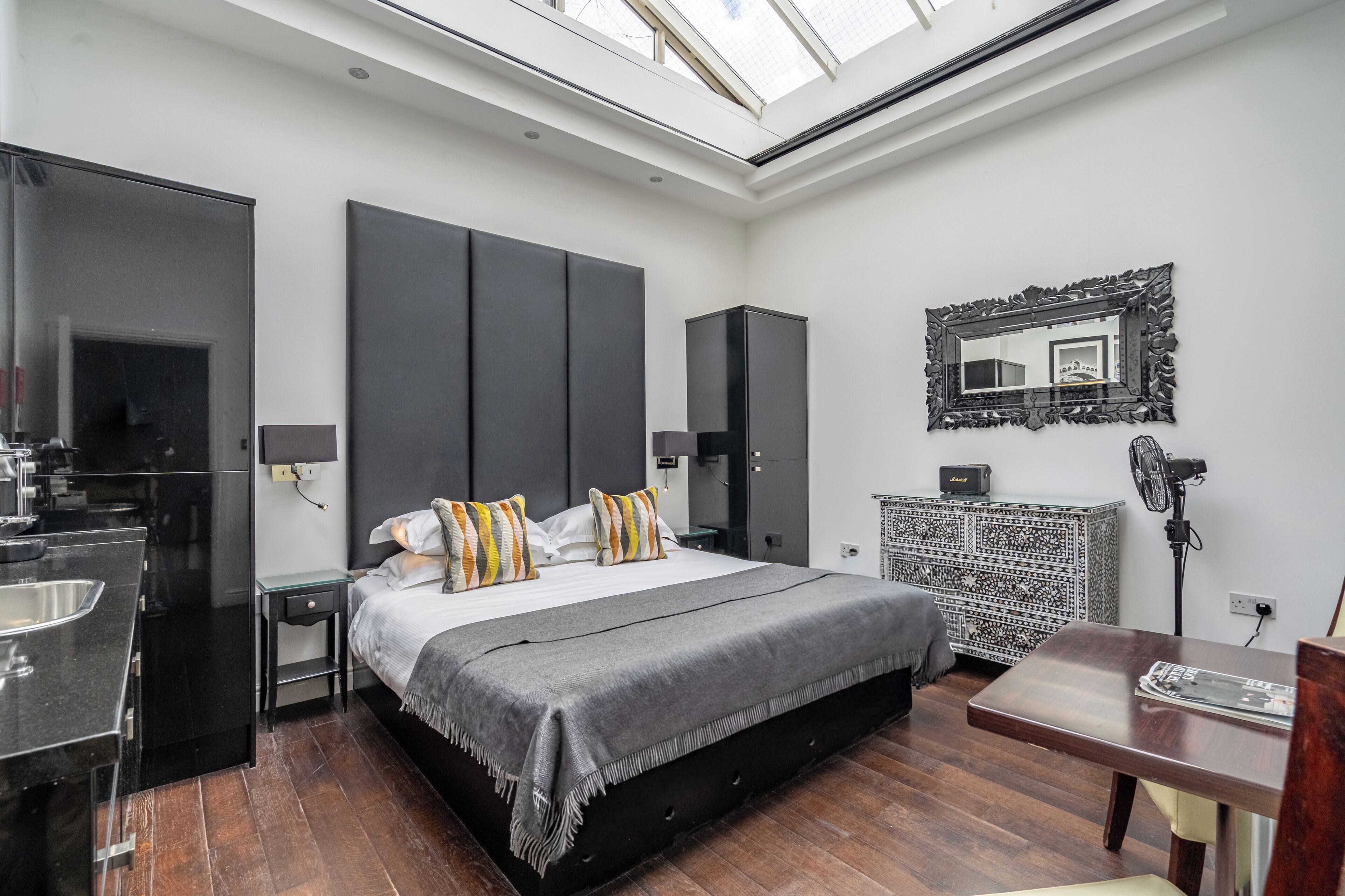 A modern bedroom with a skylight, a large bed with striped pillows, a dark wardrobe, and a small bedside table.