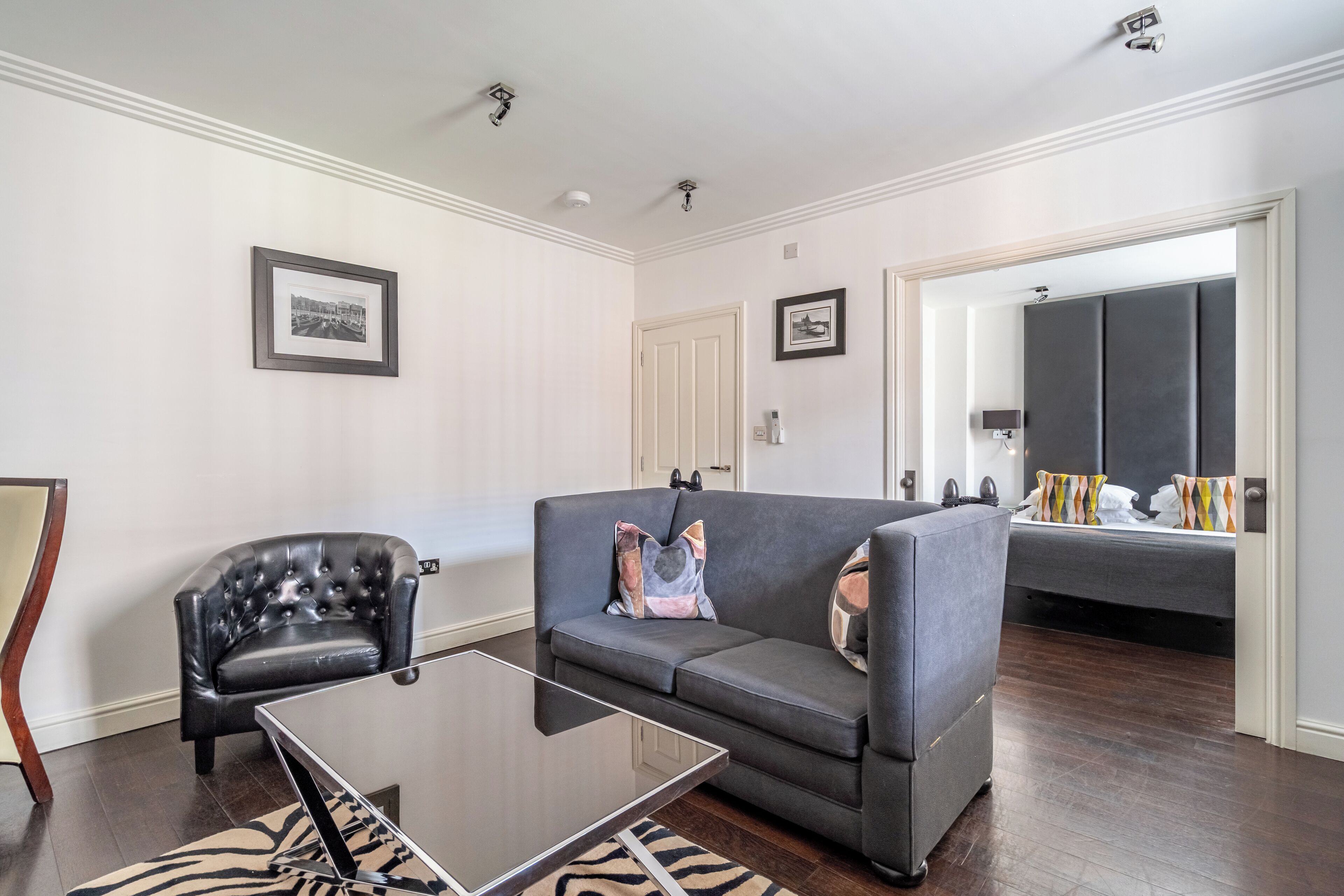 Een moderne woonkamer met een grijze bank, een zwarte leren fauteuil en een glazen salontafel. Er is een deur die leidt naar een slaapkamer met een bed en een grote spiegel.