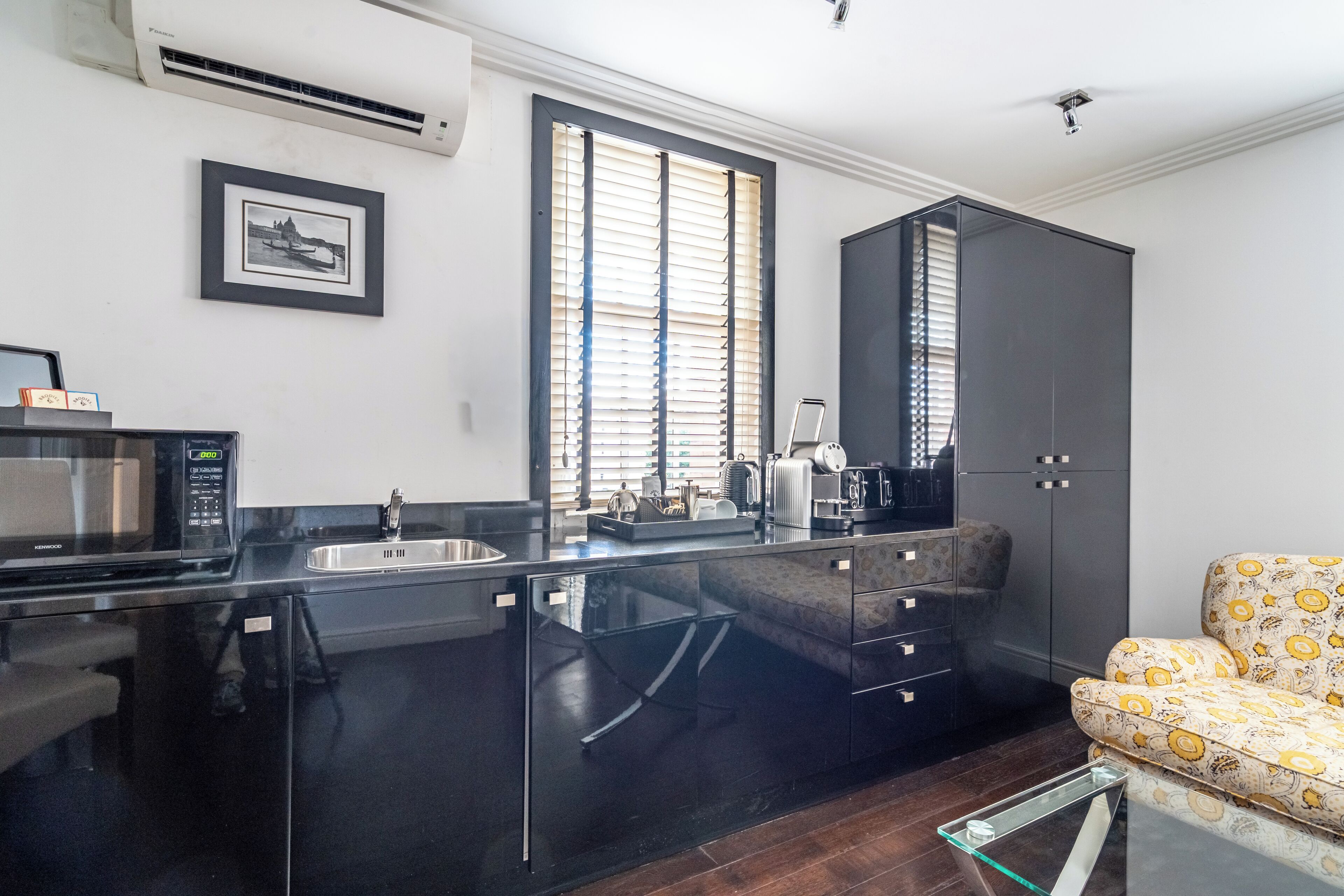 A modern kitchen with a black countertop, a sink, and a microwave. There is a window with blinds, a cabinet, and a chair with a patterned cushion.