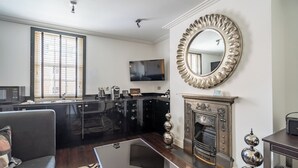 Una sala de estar moderna con chimenea, un espejo grande y una zona de cocina con armarios negros y un televisor de pantalla plana.