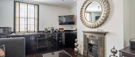 A modern living room with a fireplace, a large mirror, and a kitchen area with black cabinets and a flat-screen TV.