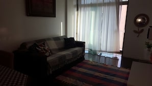 A living room with a sofa, a patterned rug, and a window with sheer curtains. - Etours - Lindo 2 Quartos no Coração de Ipanema (Rio de Janeiro)