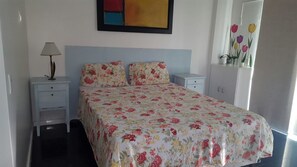 A bedroom with a bed, nightstands, a lamp, a cabinet, and a wall with a painting and flowers. - Etours - Lindo 2 Quartos no Coração de Ipanema (Rio de Janeiro)