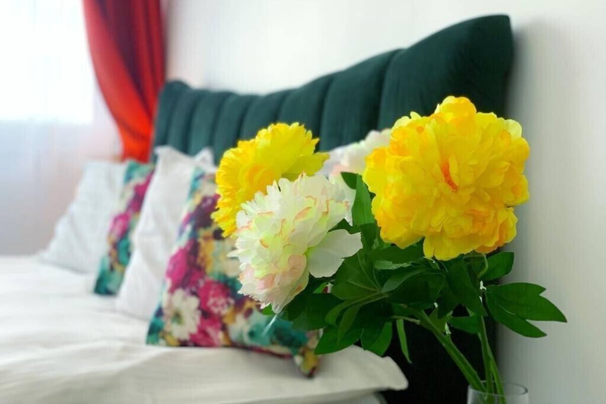 Una cama con ropa blanca y una almohada verde, un florero con flores amarillas y blancas, y una cortina roja al fondo.