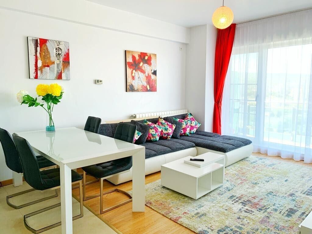 A modern living room with a white sofa, black chairs, a white coffee table, and a colorful rug. There are two paintings on the wall and a vase with yellow flowers on the table.