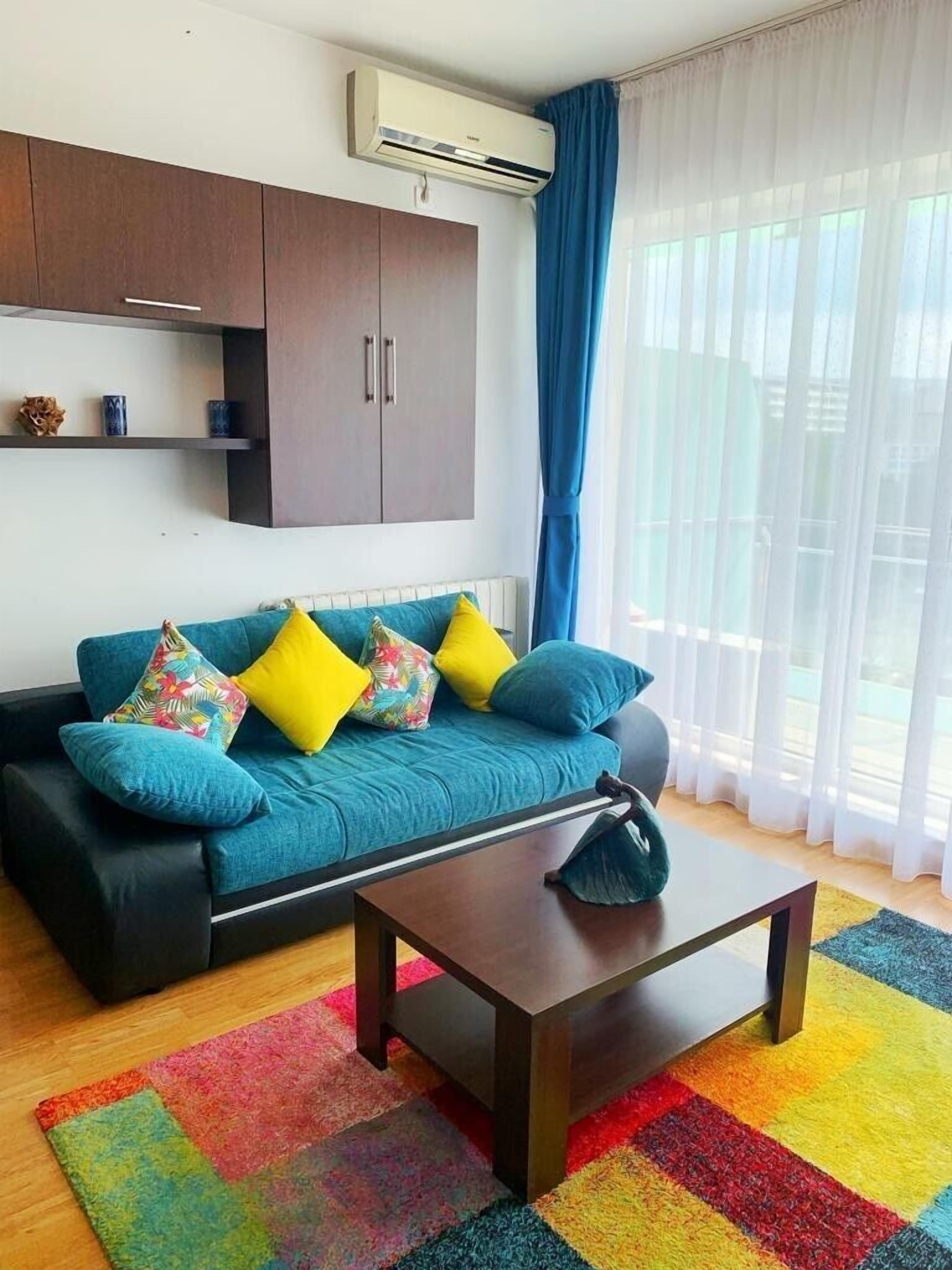 Uma sala de estar moderna com um sofá verde-água, almofadas coloridas, uma mesa de centro de madeira e um tapete vibrante.