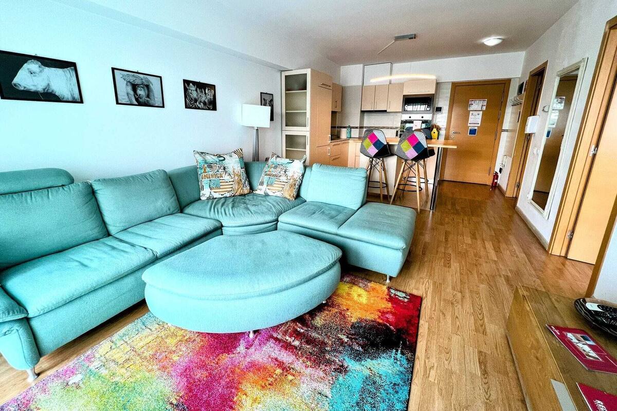 Una sala moderna con un sofá seccional verde azulado, una alfombra colorida y una cocina al fondo.