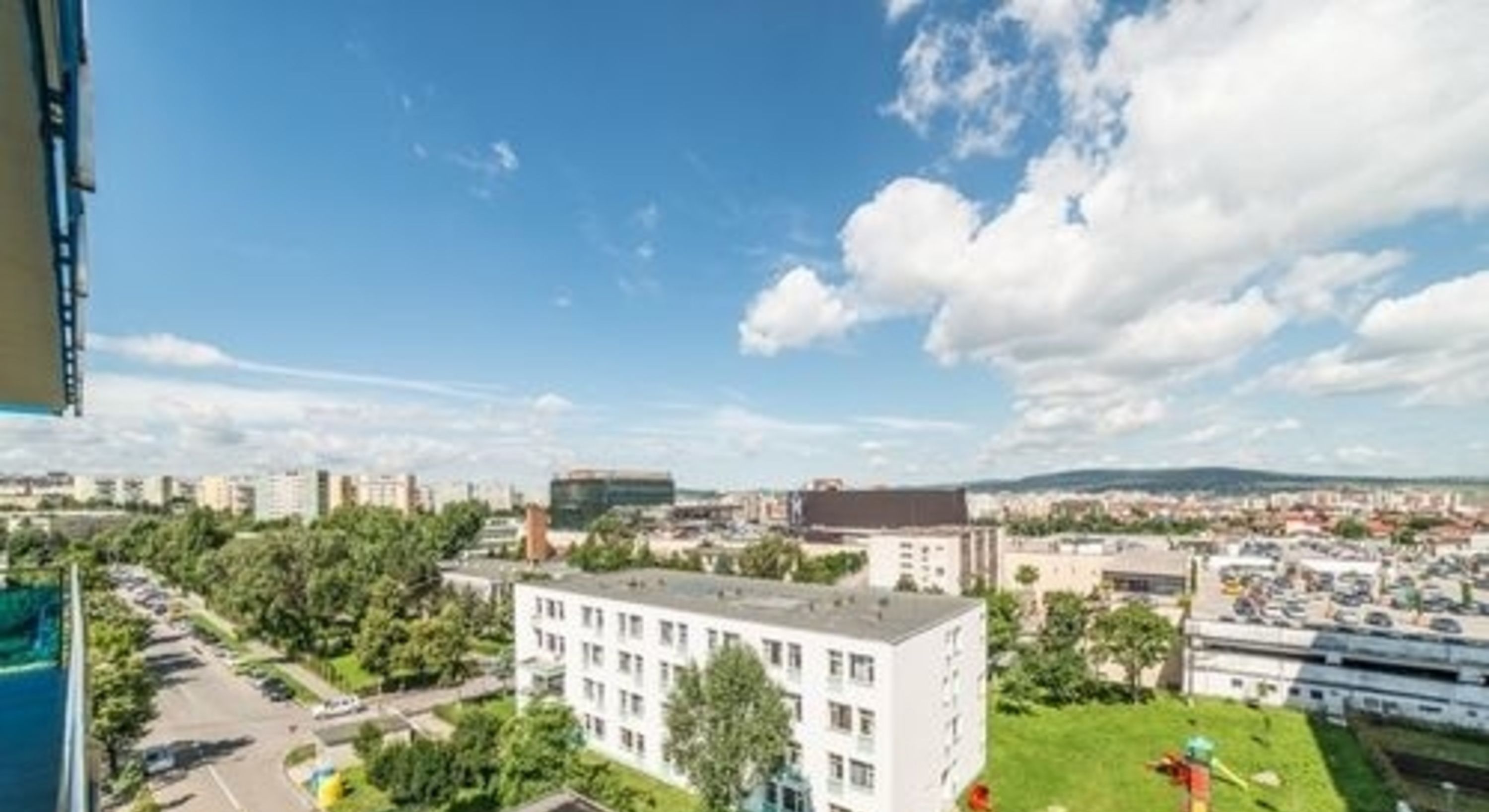 Une vue urbaine avec un grand bâtiment blanc bien visible au premier plan, entouré de verdure et sous un ciel bleu dégagé.