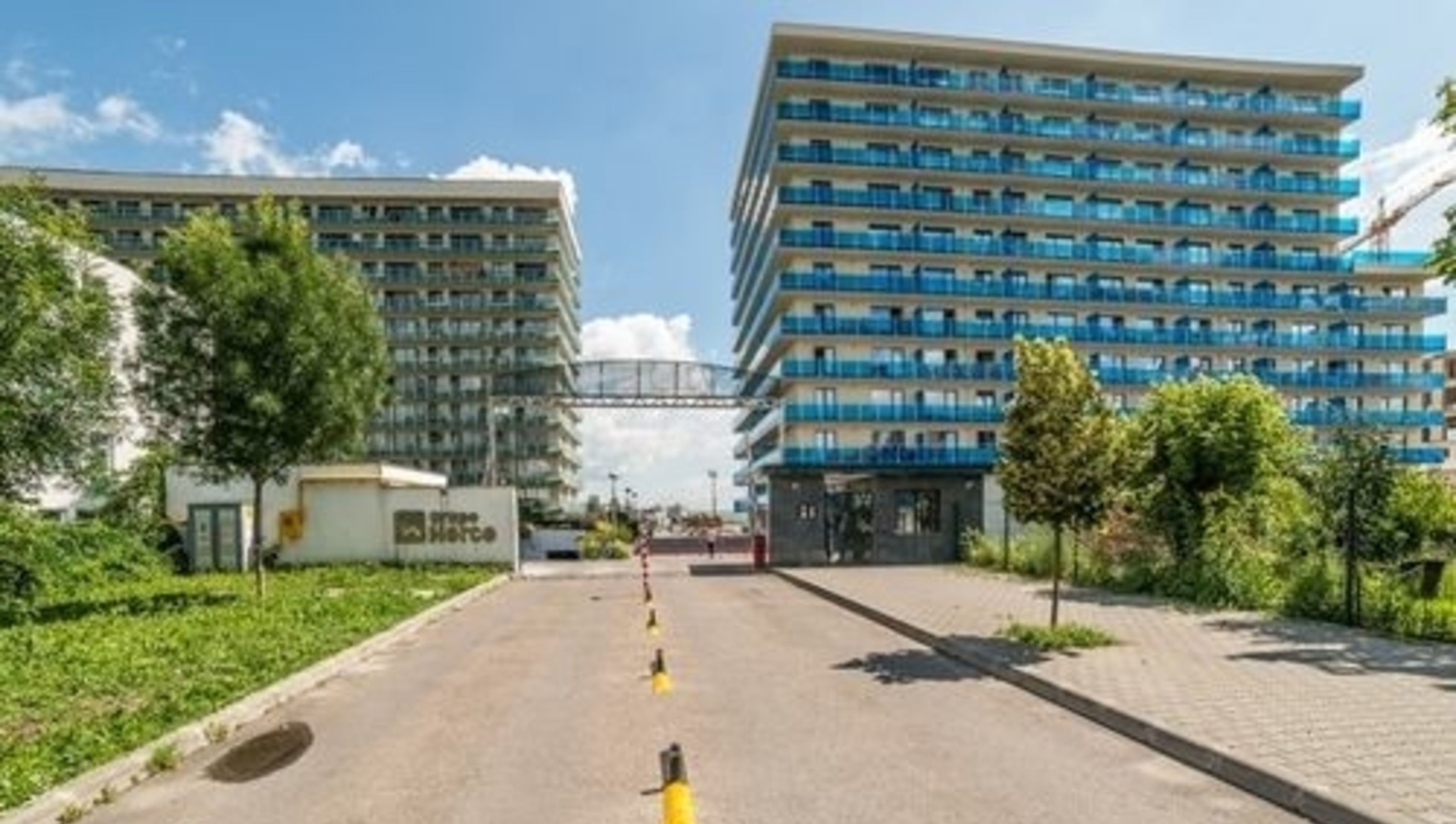 Un bâtiment moderne à plusieurs étages, doté de balcons et entouré de verdure et d’une zone pavée.