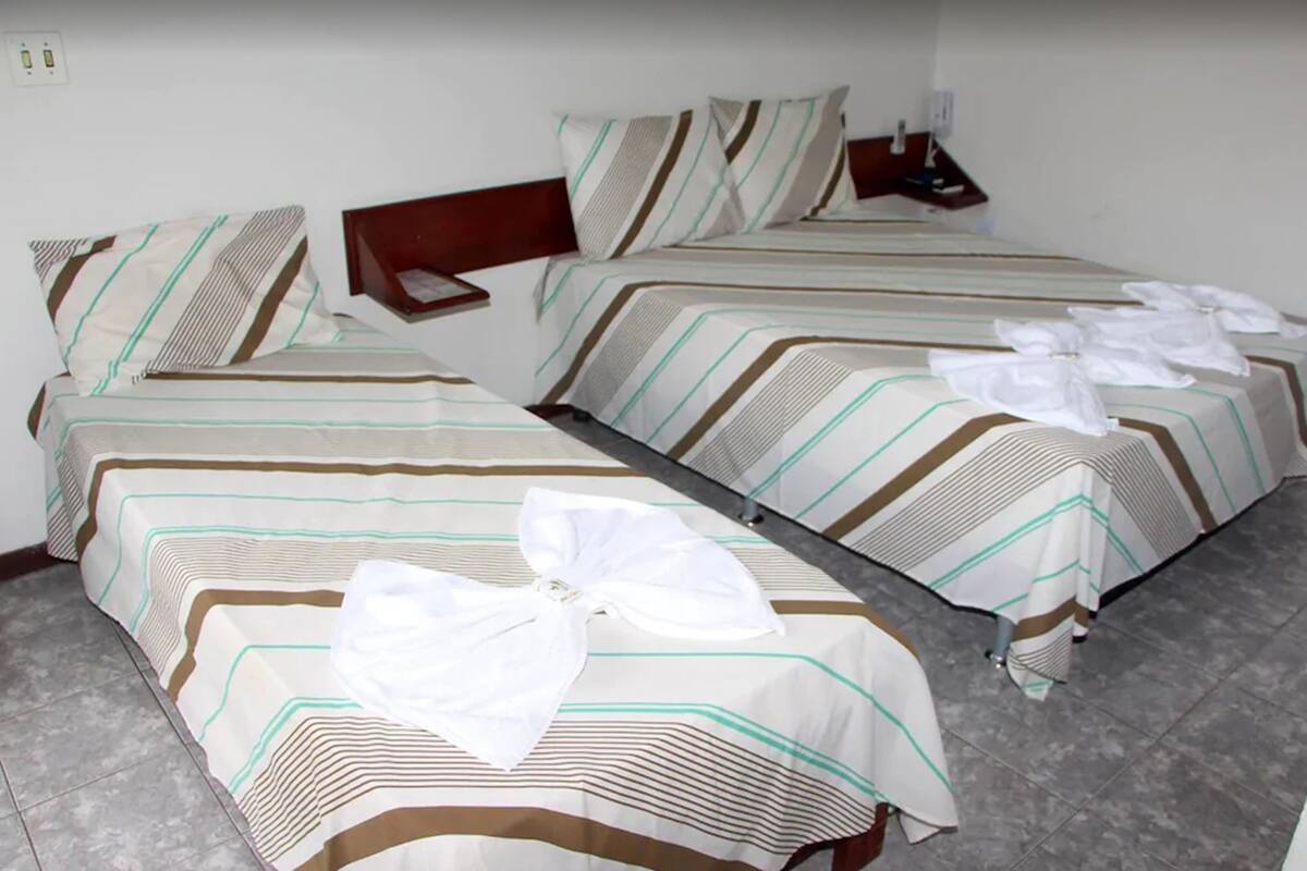 Habitación de hotel con dos camas, cada una con ropa de cama a rayas y una almohada. Hay toallas sobre las camas y una mesita de noche con una lámpara.