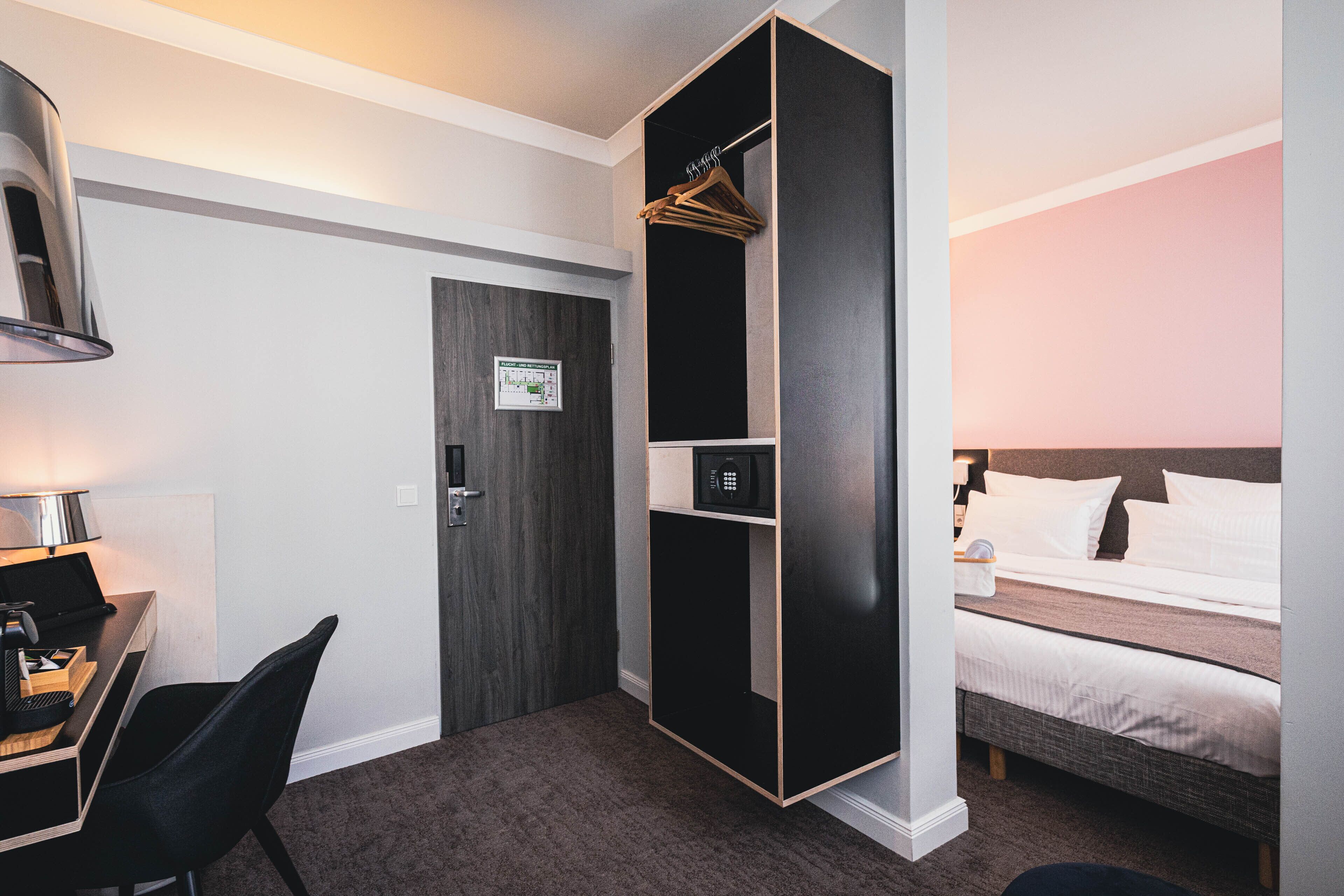 A hotel room with a desk, chair, wardrobe, bed, and a wall with a clock.
