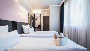 A hotel room with two beds, a bedside table with a lamp, a window with curtains, and a door leading to a bathroom. - Mountainpark Kassel (Kassel)