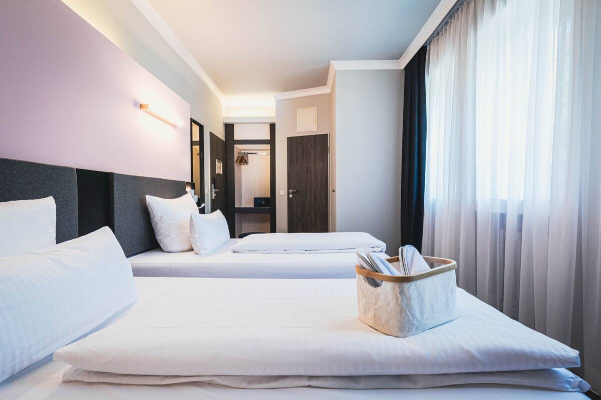 A hotel room with two beds, a bedside table with a lamp, a window with curtains, and a door leading to a bathroom.