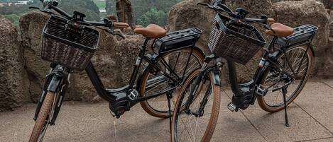 Deux vélos, équipés de paniers en osier et de selles en cuir marron, sont garés sur une surface en pierre, avec une vue sur la ville en arrière-plan.