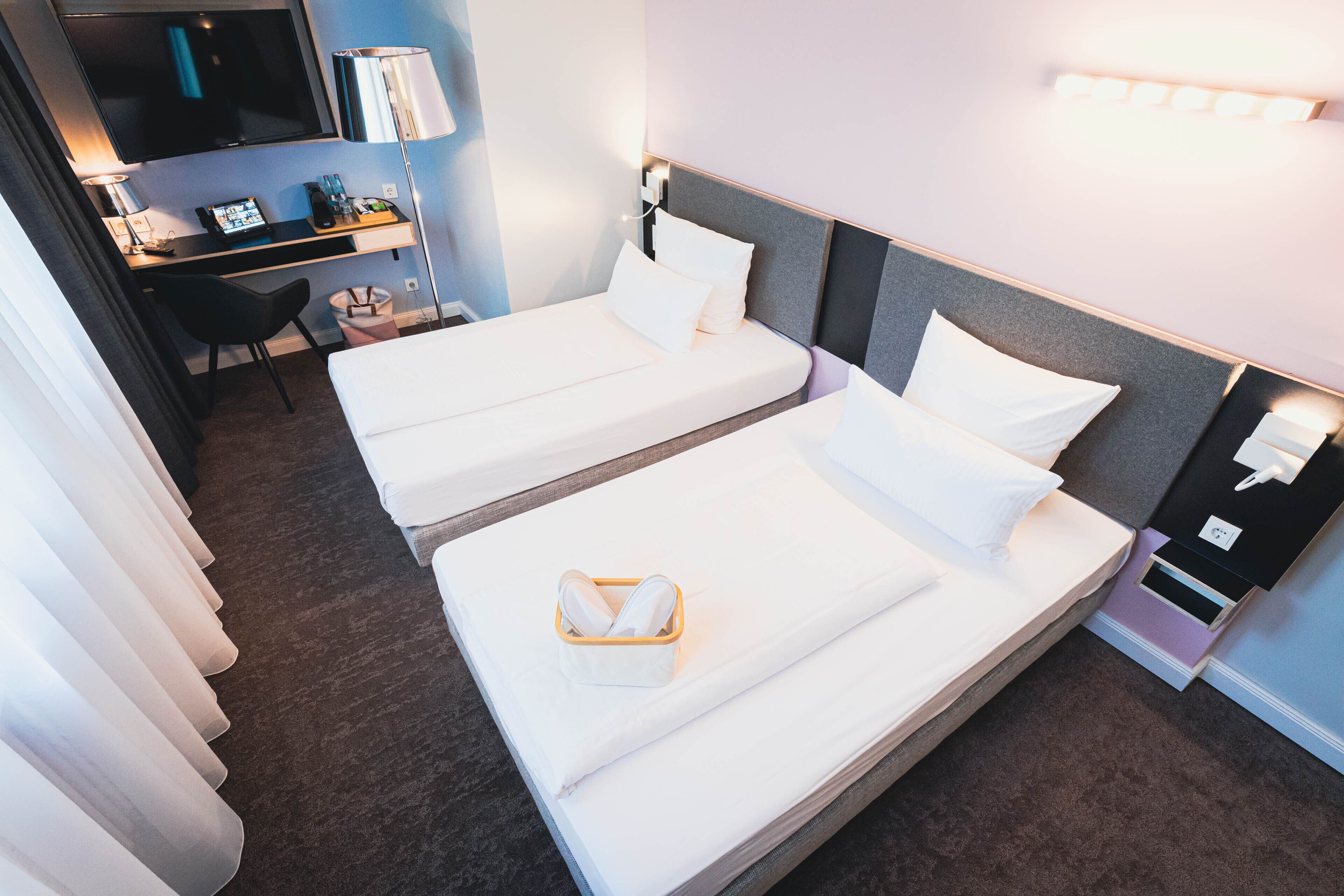 A hotel room with two beds, a desk with a computer, a TV, and a bedside table with a lamp.